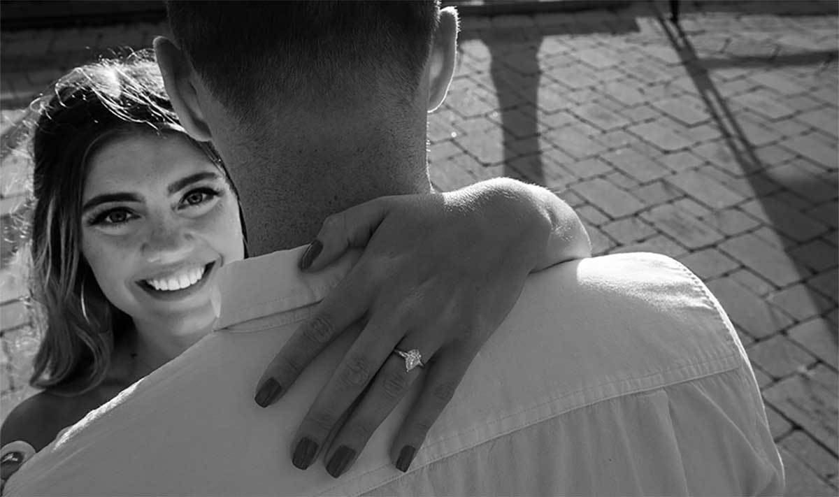 A woman smiles at the camera while embracing a man, displaying an engagement ring on her left hand.