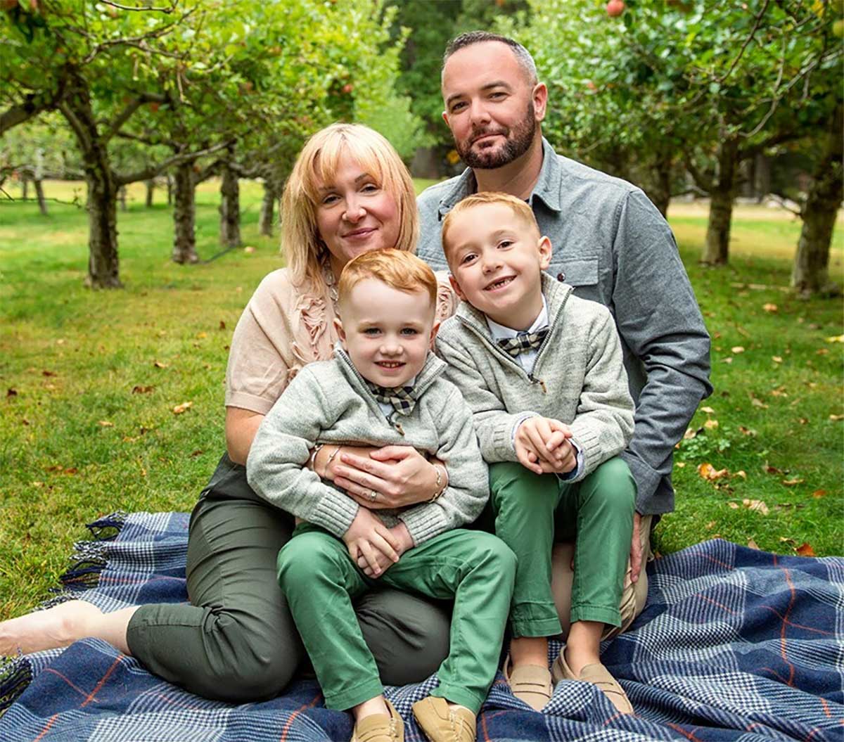 Four people, two adults and two young boys, sit on a blanket in an orchard, posing for a family photo surrounded by green grass and trees.