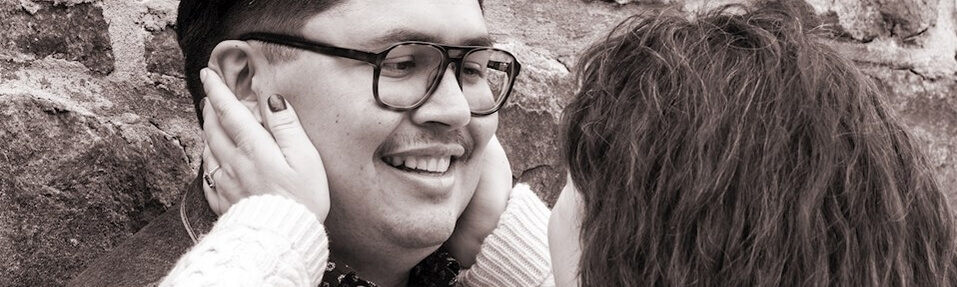 A person with glasses smiles as another person holds their face with both hands near a brick wall.