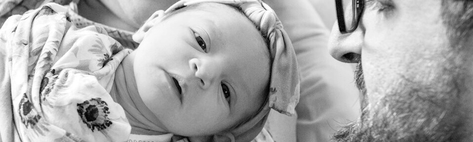 A newborn baby wrapped in a patterned blanket looks up while being held by an adult, with another adult’s face visible nearby.