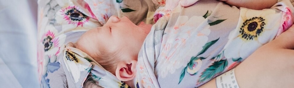 A newborn baby wrapped in a floral-patterned blanket and headband is held by an adult wearing matching floral clothing and a hospital wristband.