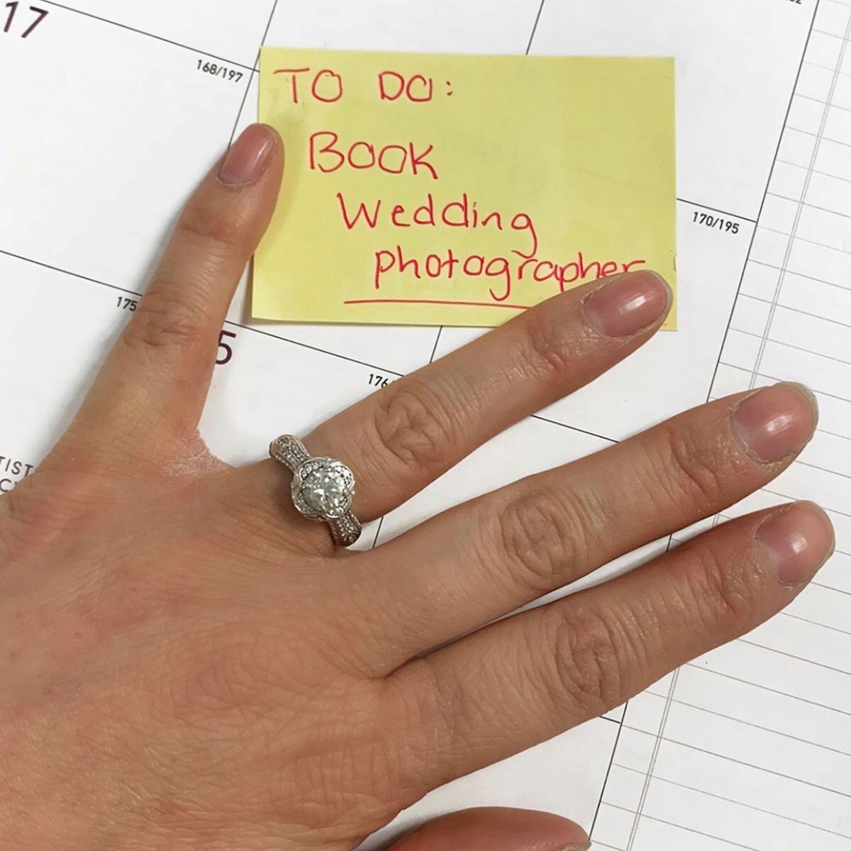 A hand with an engagement ring rests on a calendar next to a sticky note that says, "TO DO: Book wedding photographer.
