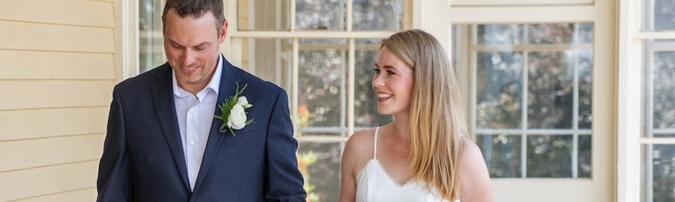 A man in a suit with a white rose boutonniere stands next to a woman in a white dress, both smiling indoors near large windows.