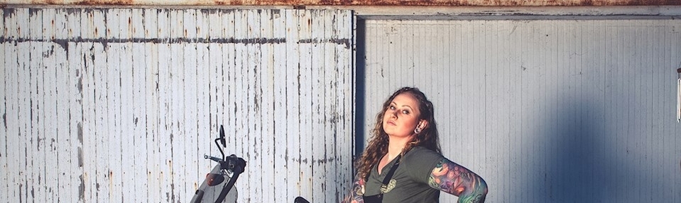 A woman with tattooed arms stands next to a motorcycle in front of a weathered, rusted metal door.