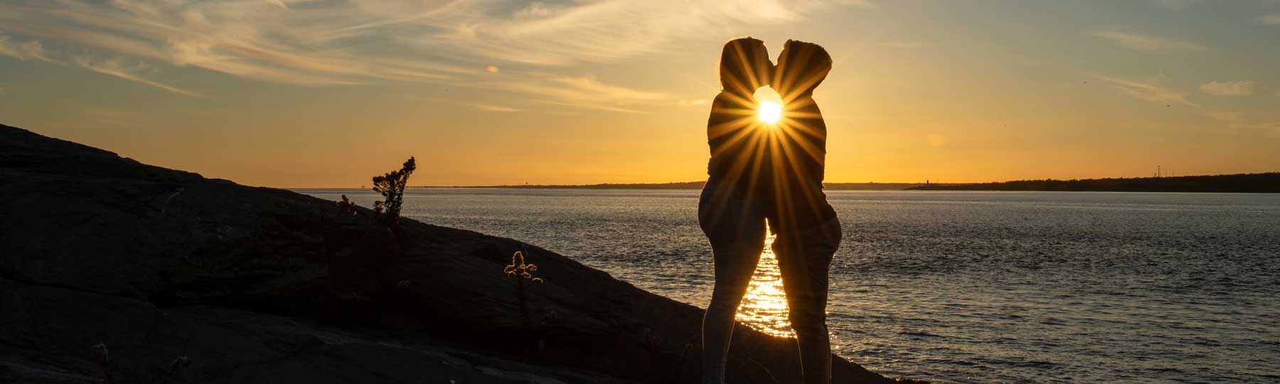 Two people stand close together on a rocky shore at sunset, silhouetted against the sky with the sun shining between them over the water.