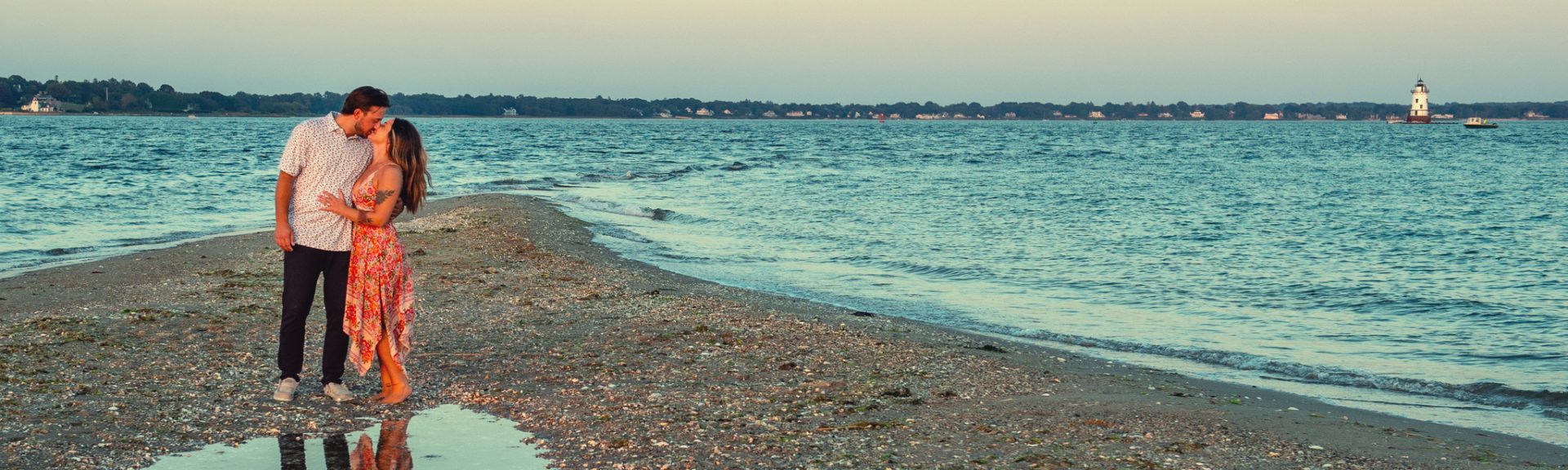 A couple stands close together and kisses on a narrow stretch of beach with the ocean and a distant lighthouse in the background.