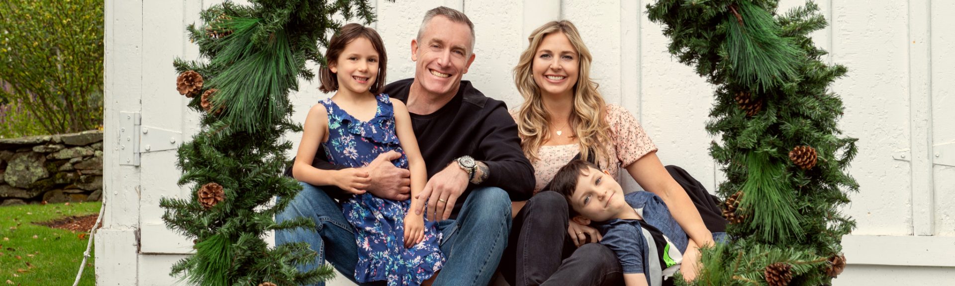 A family of four sits together outdoors, framed by two large holiday wreaths, posing and smiling in front of a white wooden wall.