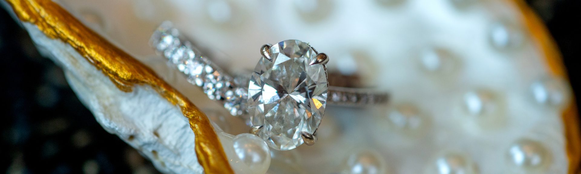 An oval-cut diamond ring and a diamond-studded band are displayed on a pearlescent seashell with gold edges.