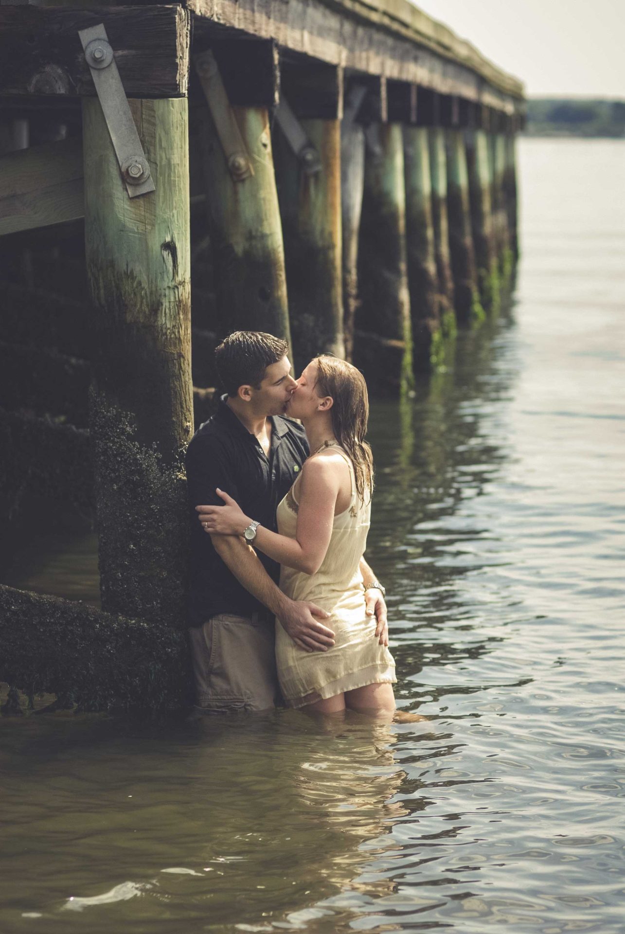 rhode-island-engagement-ocean-pier A couple stands in shallow water under a wooden pier, embracing and kissing.