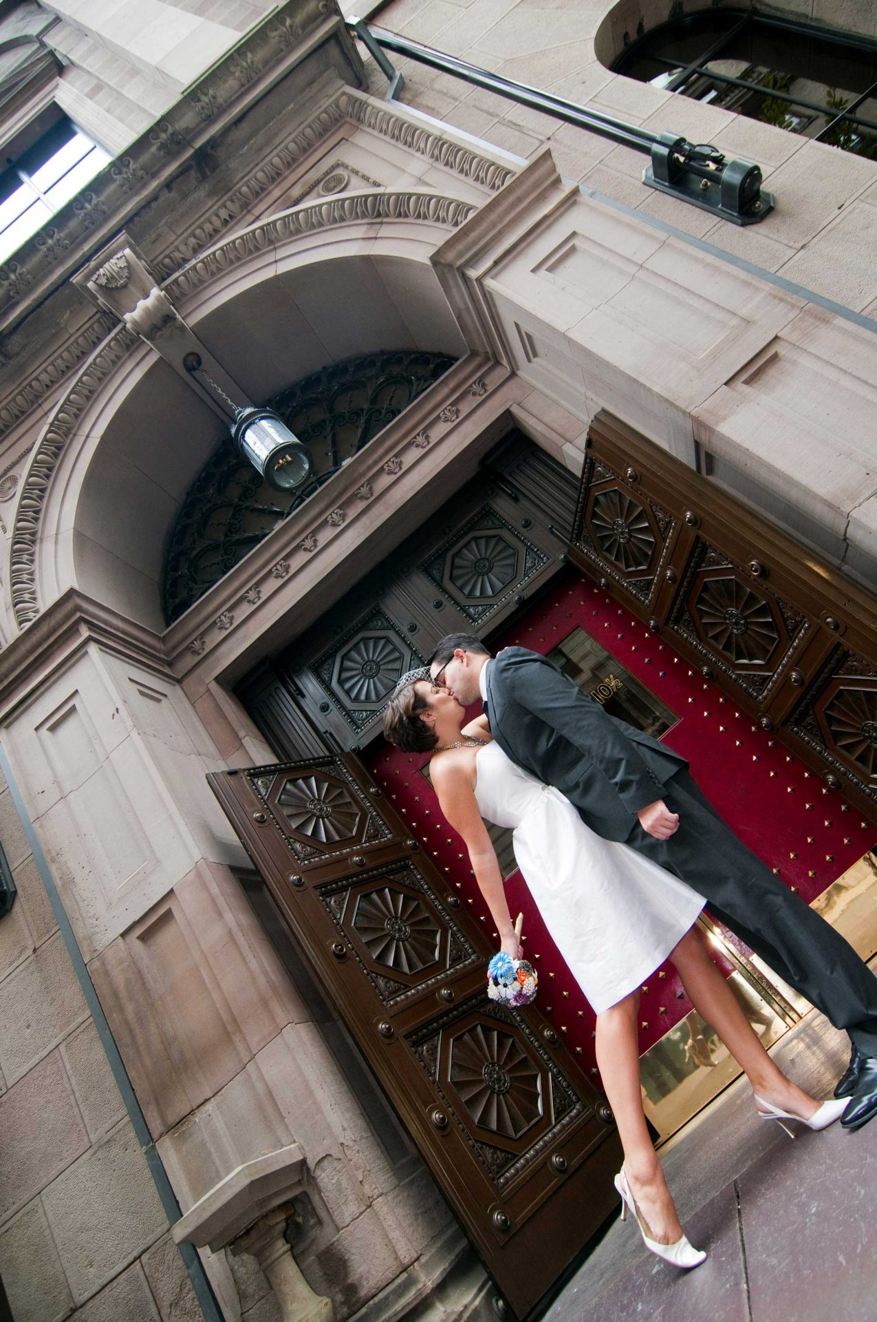 wedding-couple-in-front-of-church-angle A couple dressed in wedding attire kisses in front of a large, ornately carved wooden door on a stone building. The bride holds a bouquet of flowers.