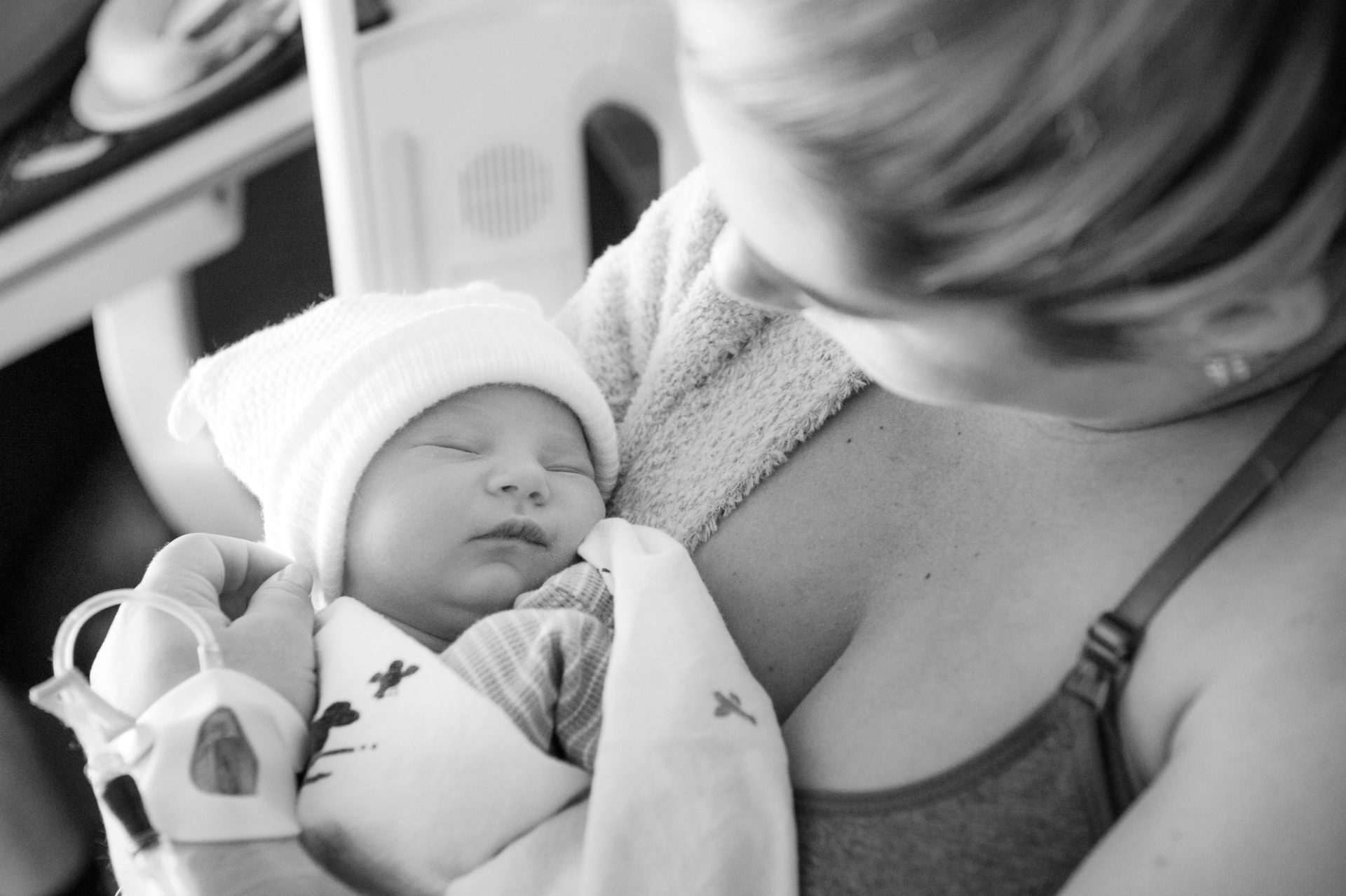 family-photo-newborn-in-hospital-bw A woman holds a newborn baby wrapped in a blanket, wearing a knit hat, in a hospital setting.