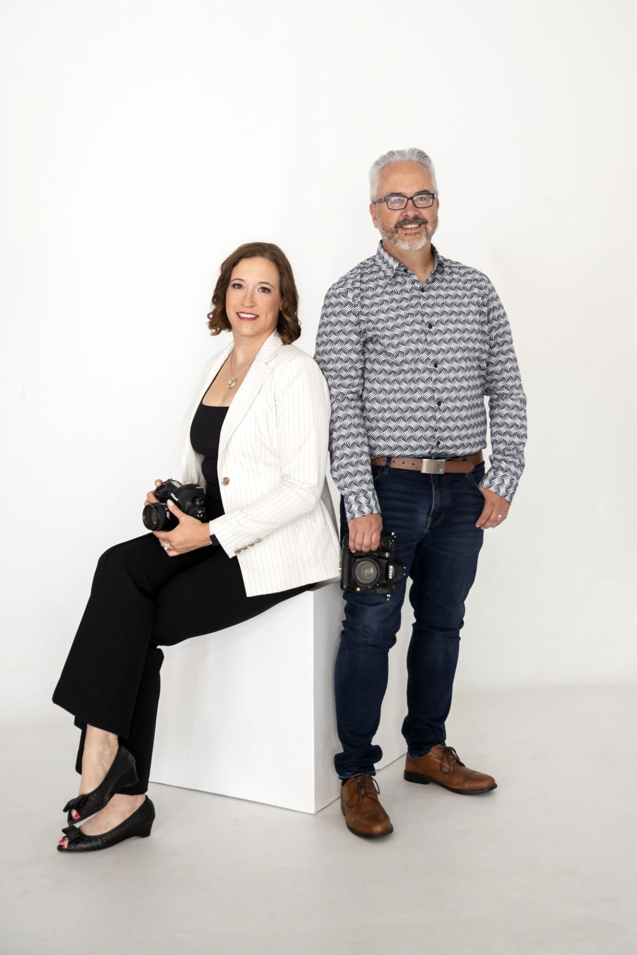 A woman in a white blazer sits holding a camera, while a man in a patterned shirt and jeans stands next to her, also holding a camera, against a white background.