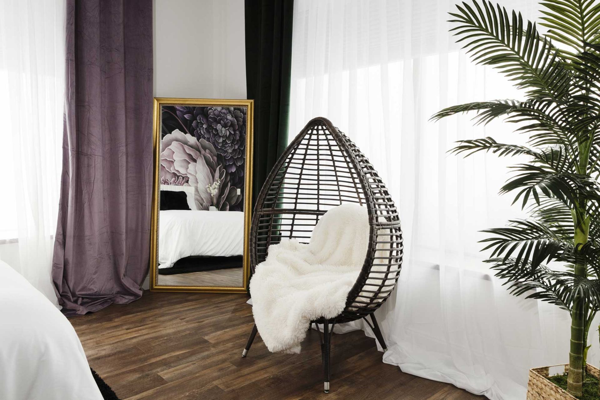 nicole-gesmondi-boudoir-seat-studio A wicker egg chair with a white throw sits by sheer curtains, next to a tall mirror and a large potted plant in a modern bedroom with wood flooring.