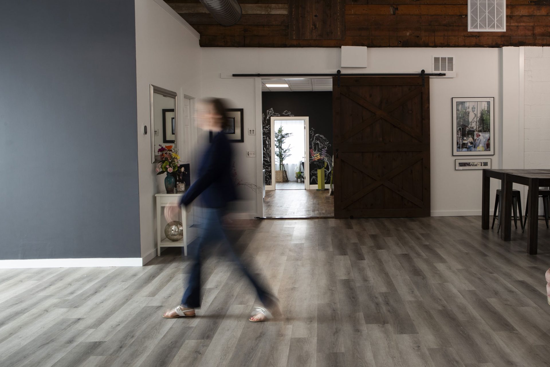 nicole-gesmondi-in-her-photography-studio A person walks through a modern room with gray flooring, a wooden sliding door, wall art, and a table with chairs.