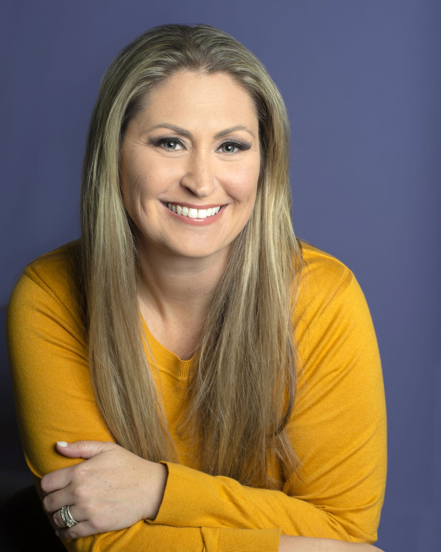 ri-headshot-girl-in-yellow-purple-bkgd A woman with long blonde hair wearing a yellow sweater smiles at the camera against a plain purple background.