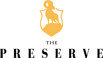 A gold silhouette of a ram standing on a hill inside a shield above the words “The Preserve” in bold black letters.