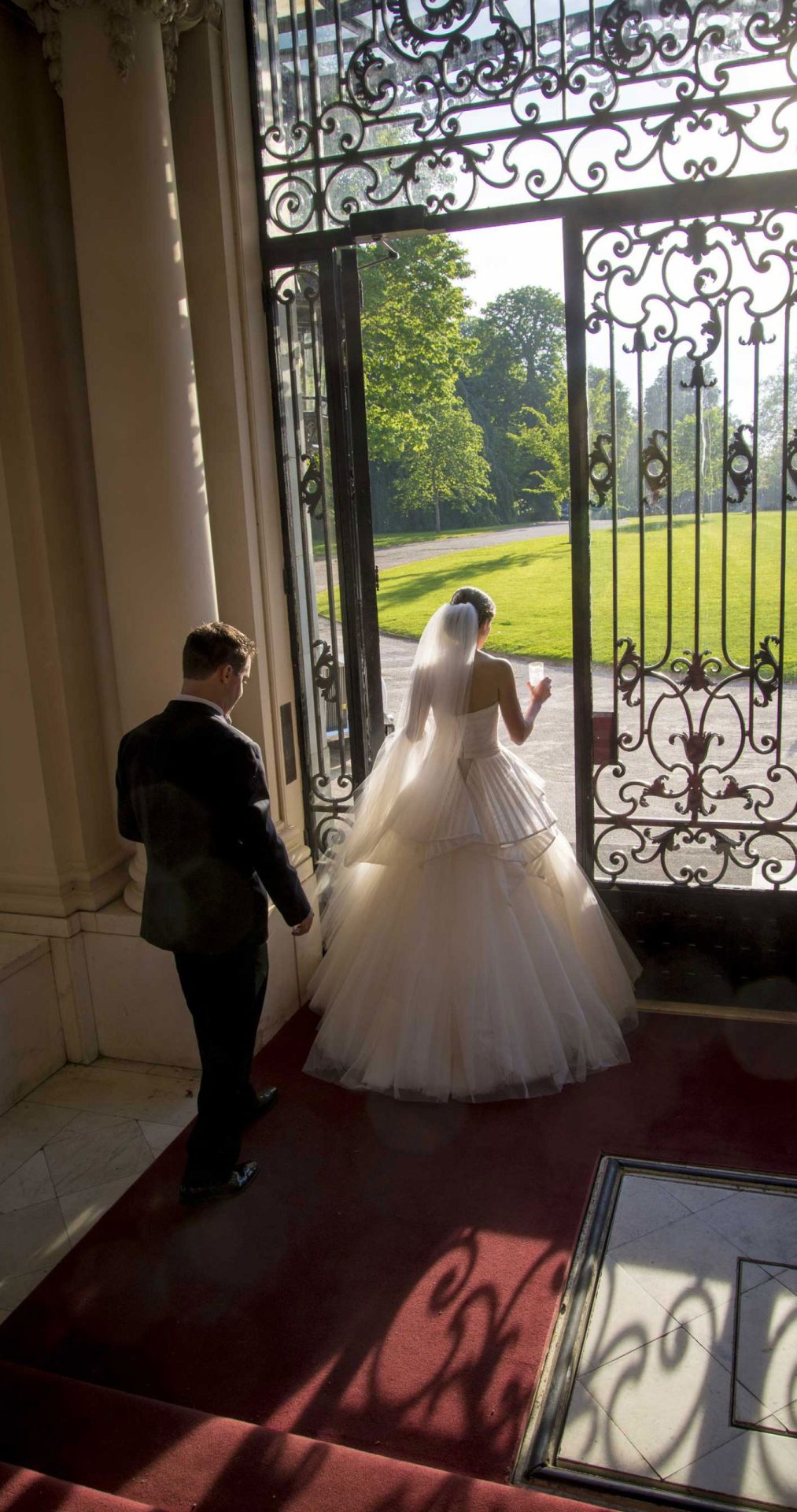 wedding-at-rosecliff-in-newport-front-door-bride-and-groom-trim A bride in a white gown and a groom in a black suit stand at ornate open doors, sunlight streaming in, looking outside onto a green lawn.