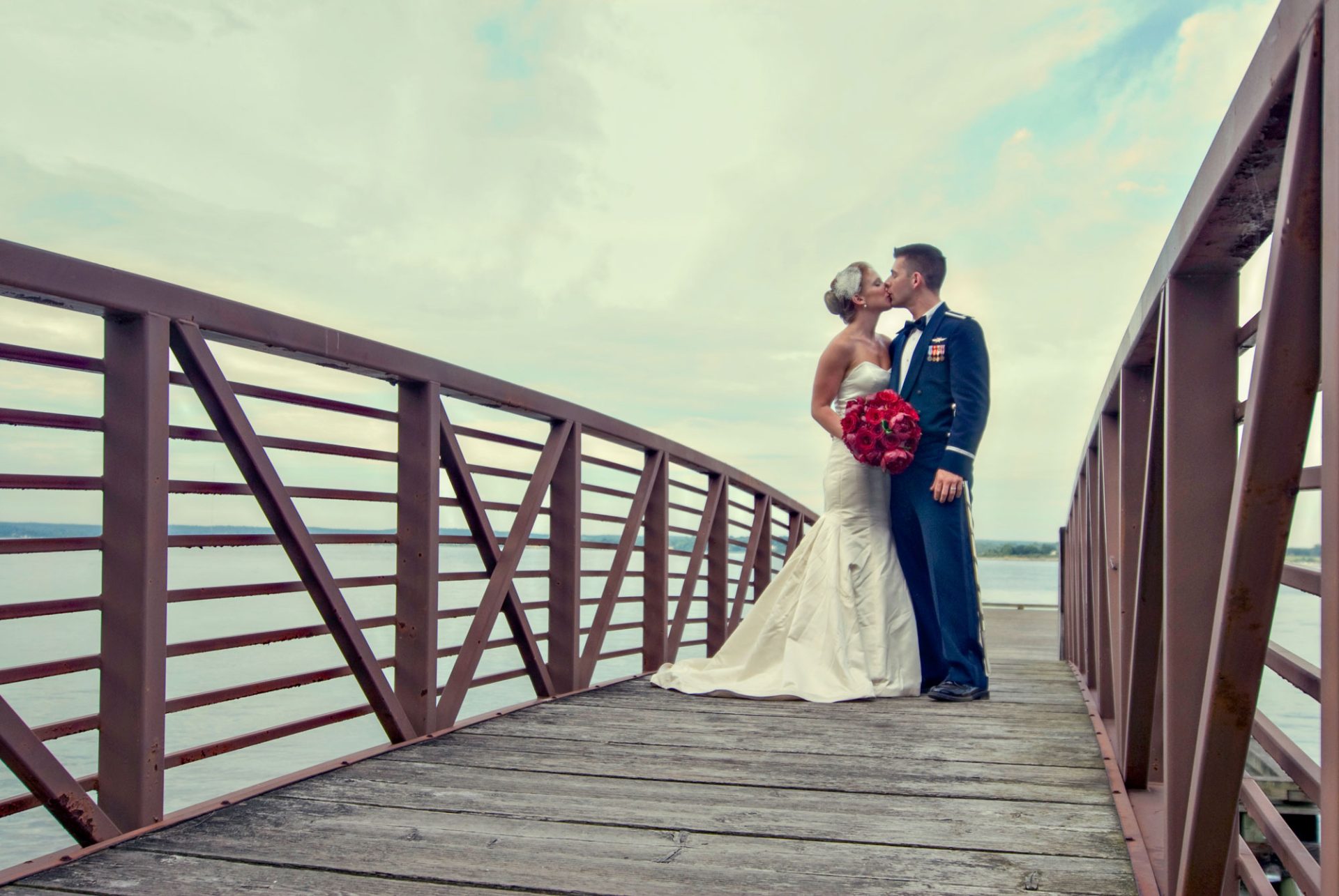 A bride in a white dress and a groom in a military uniform kiss on a wooden bridge over water under a cloudy sky.