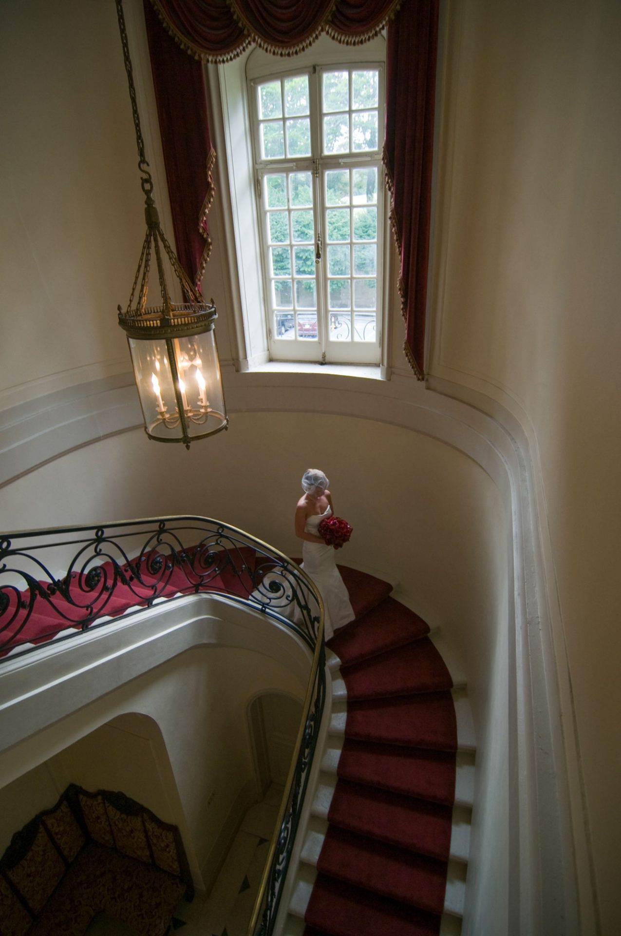 A bride in a white dress holding red flowers walks up a curved staircase with a red carpet in a building with tall windows and an ornate chandelier.