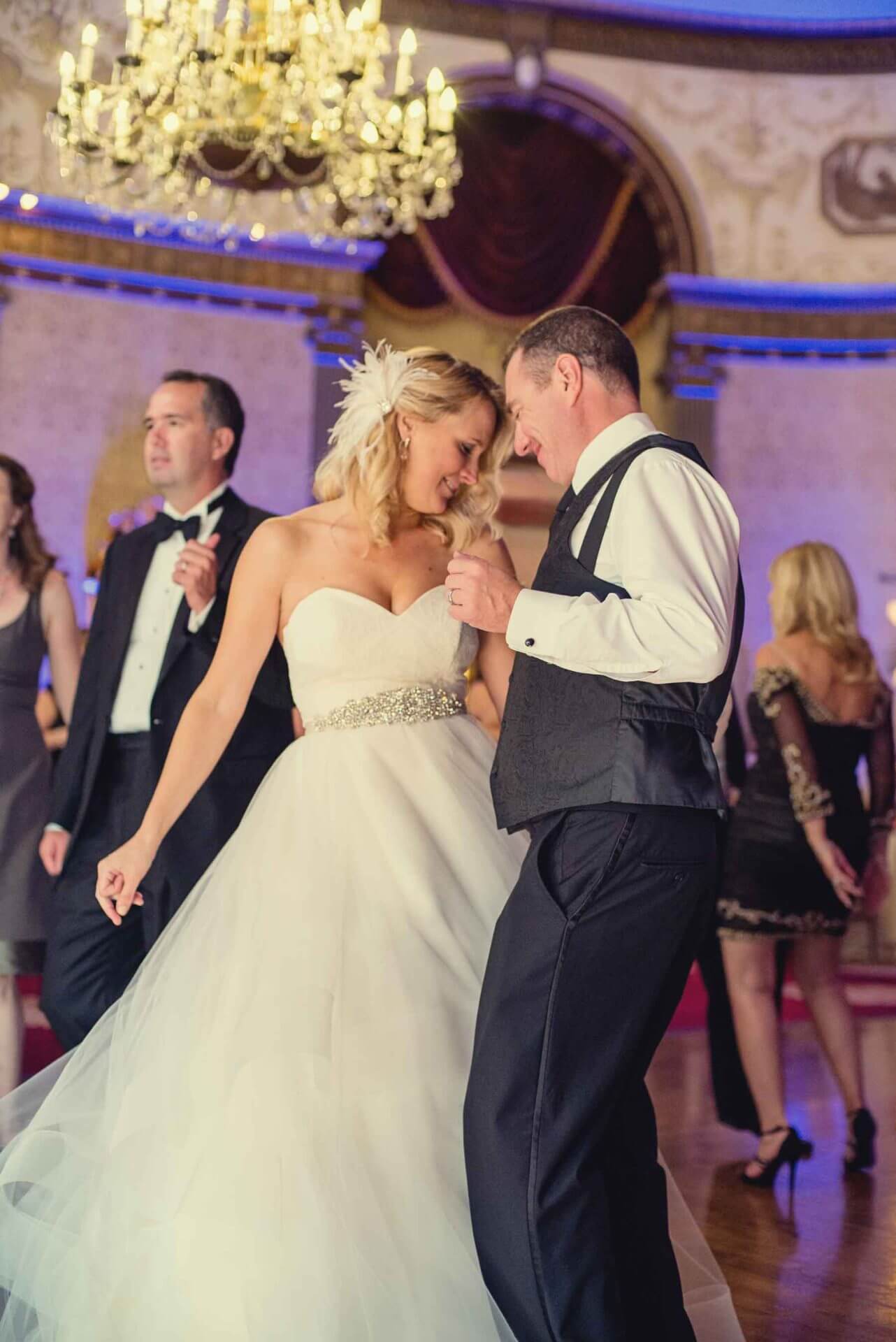 A bride and groom dance together at their wedding reception in an elegant ballroom, with other guests visible in the background.