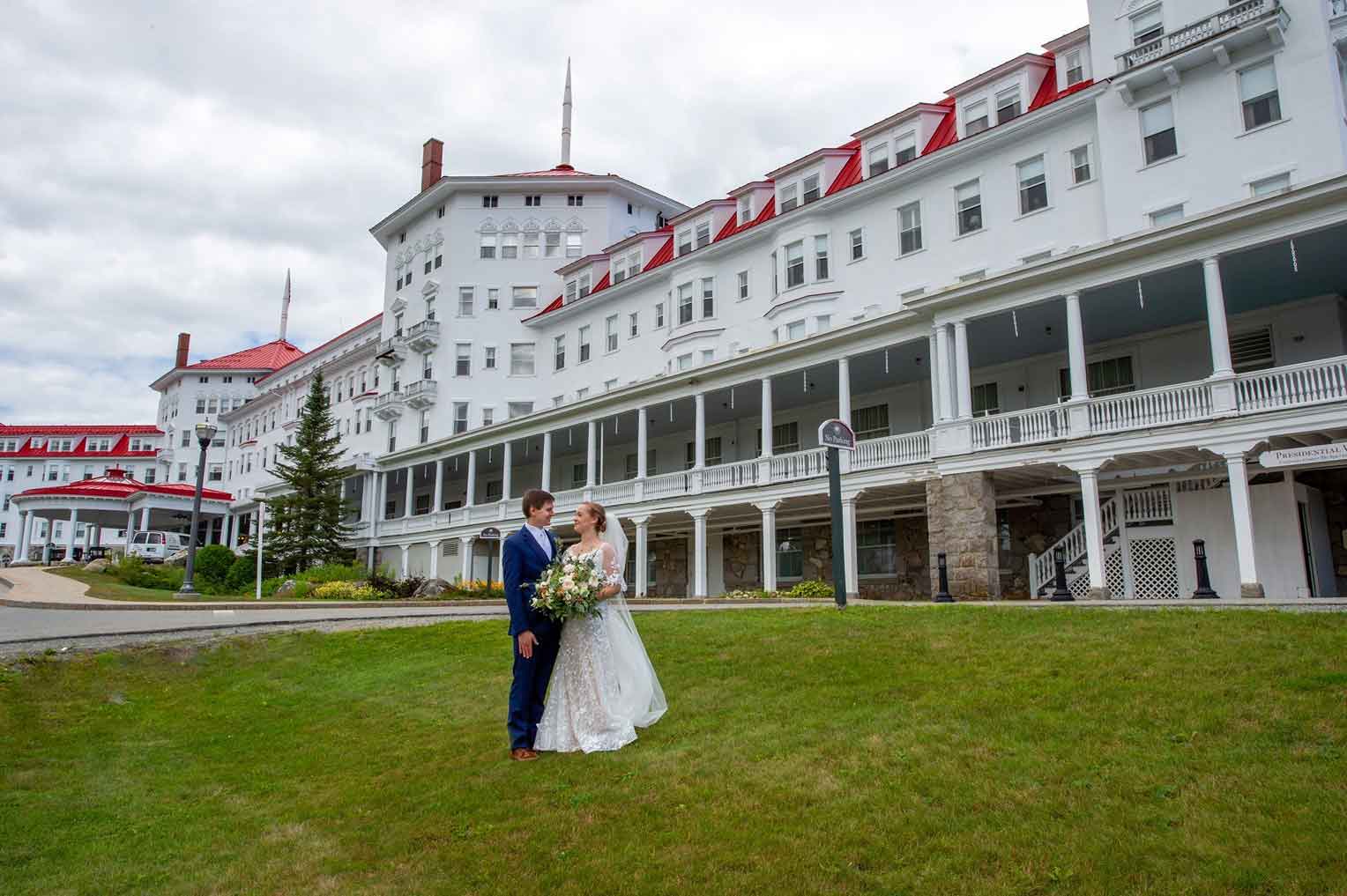 A bride and groom stand on the lawn in front of a large white historic hotel with red roofs under a cloudy sky.