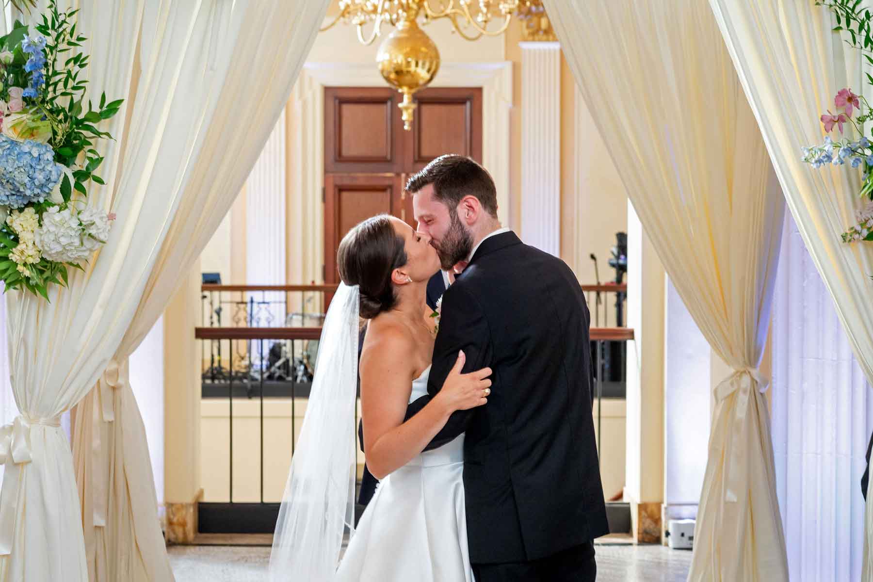 A bride and groom kiss under a floral archway at an indoor wedding ceremony, with cream-colored drapes and a chandelier overhead.