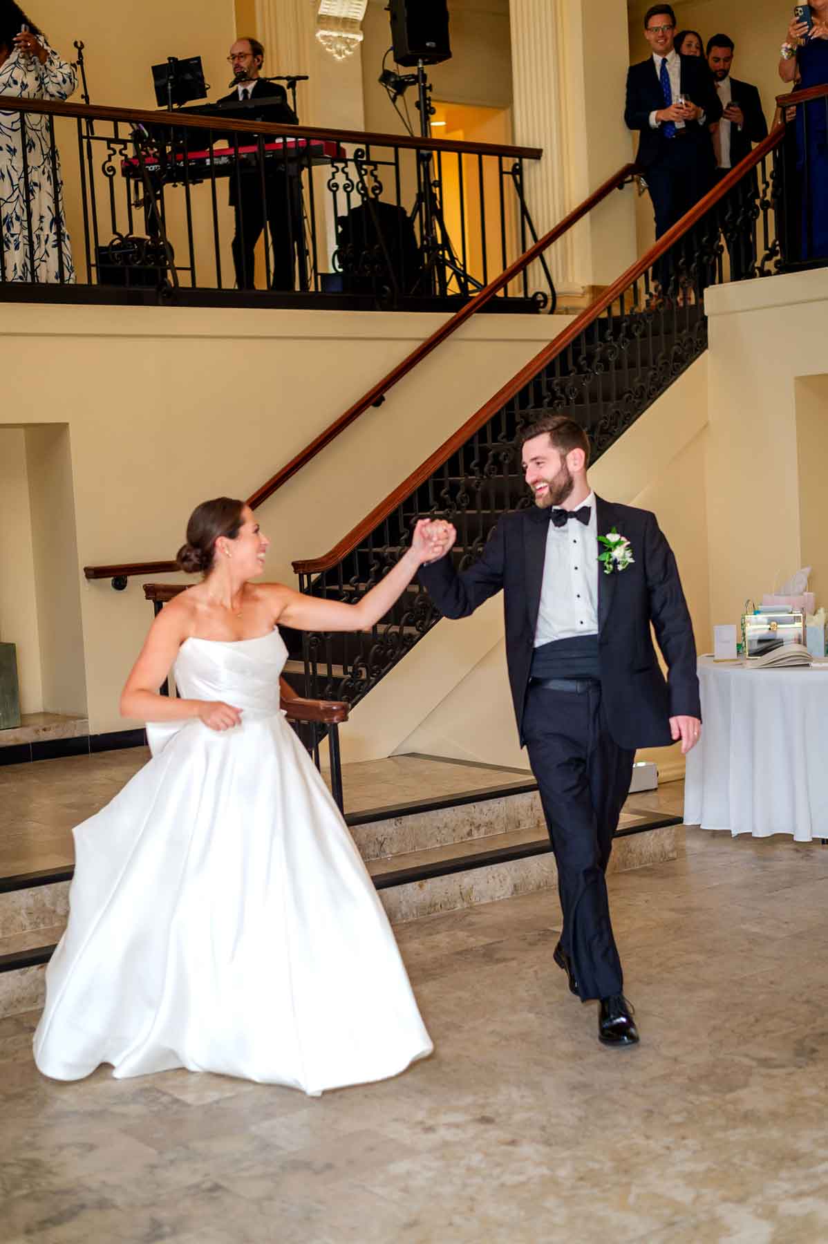 Providence-Public-Library-Wedding_68-entrance A bride and groom, both smiling, dance together indoors while wedding guests watch from a balcony and the ground floor.