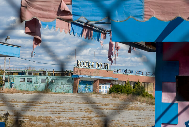 Rocky-Point-Park_11 Abandoned amusement park entrance with a faded "Rocky Point" sign, peeling paint, and overgrown weeds, viewed through a chain-link fence.