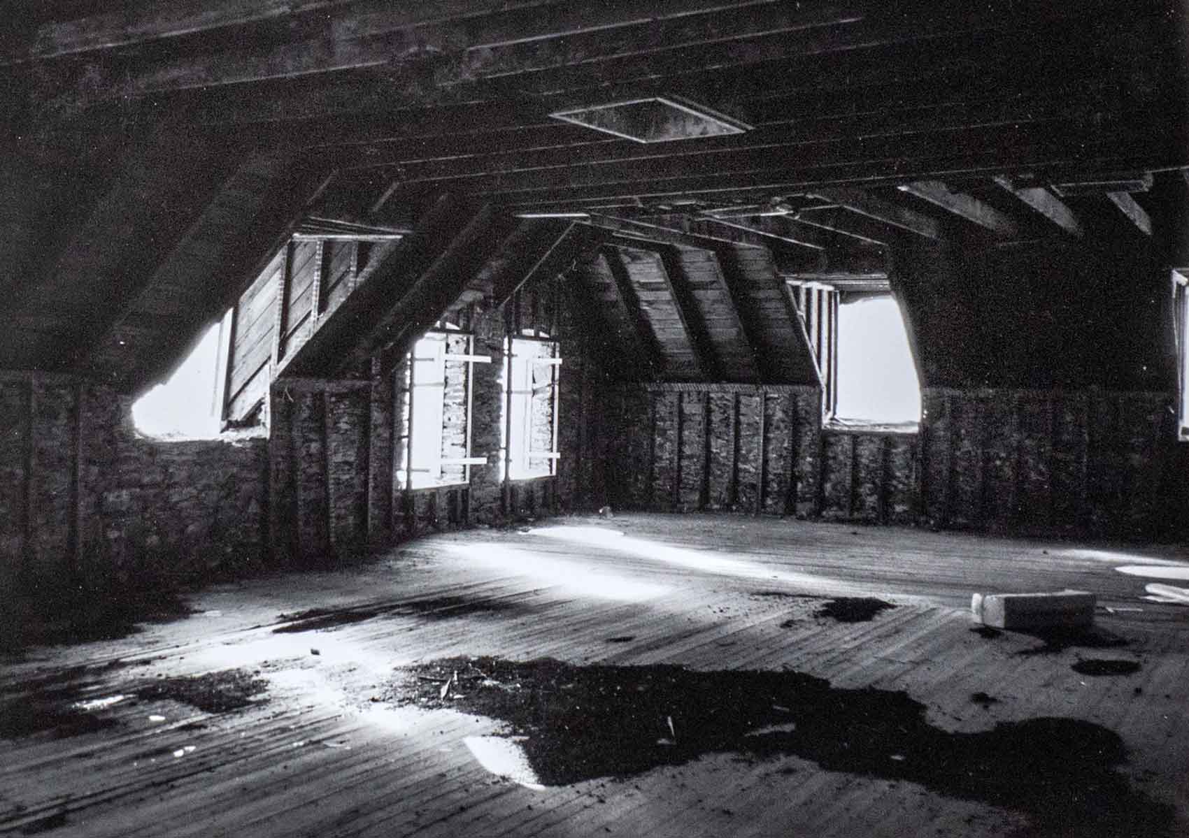 Sockanosset-Boys-Training-School-Cranston-ri Black and white photo of an empty attic with exposed beams, open windows, rough wooden floors, and patches of light and shadow.