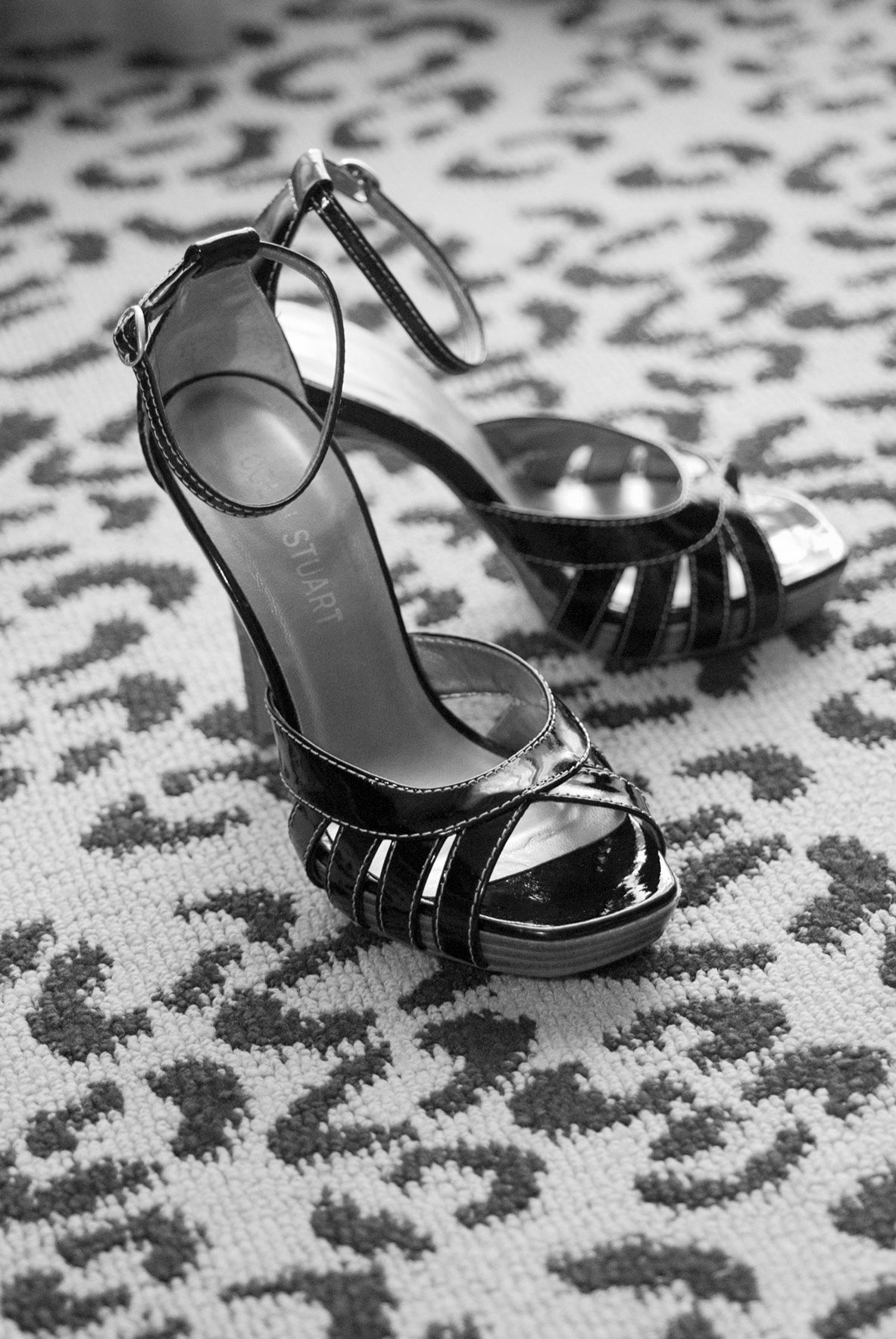 A pair of black, high-heeled, open-toe sandals with ankle straps placed on a patterned carpet.