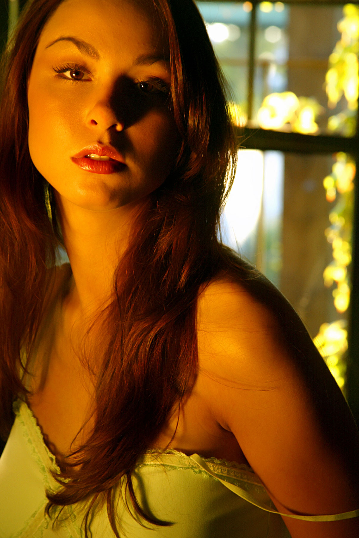 A woman with long brown hair stands indoors near a sunlit window, wearing a light-colored top and looking slightly past the camera.