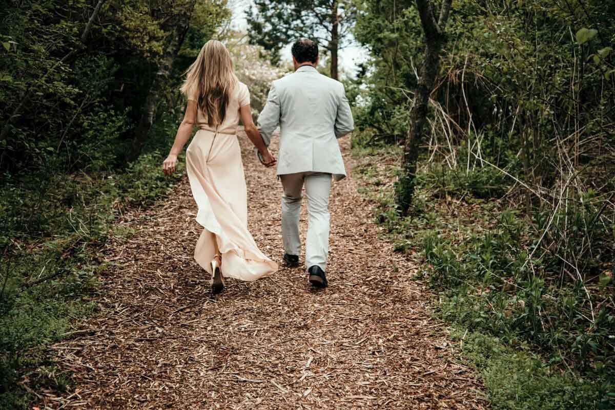 A woman in a light dress and a man in a light suit walk hand in hand up a wooded path, surrounded by greenery.