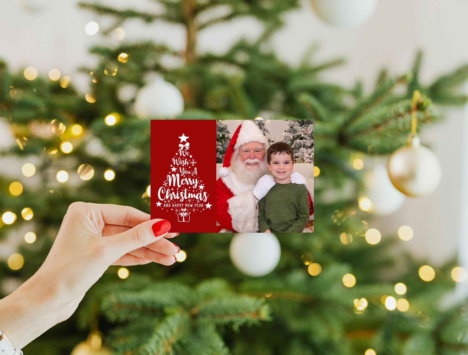 A hand holds a Christmas card featuring a photo of Santa with a young boy and festive text; a decorated Christmas tree is in the background.