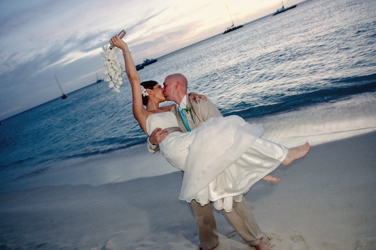 A groom lifts and kisses his bride on a sandy beach at sunset, with the bride holding a bouquet and sailboats visible in the background.