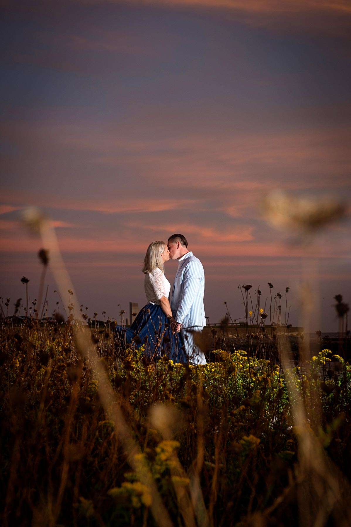 engagement-sunset-colors-kiss A couple stands close together in a field of wildflowers at sunset, facing each other under a pink and purple sky.