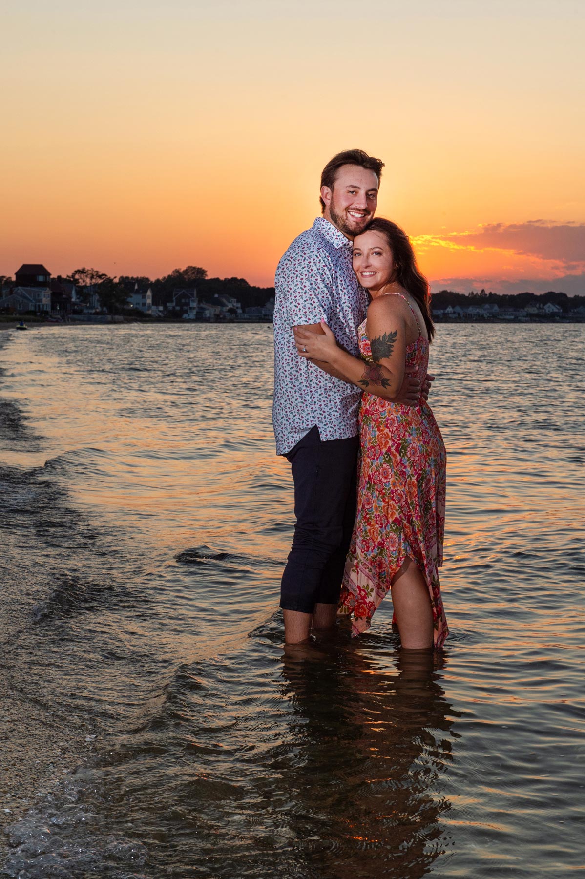 engagement-sunset A couple stands in shallow water at the beach, embracing and smiling, with a sunset and distant shoreline in the background.