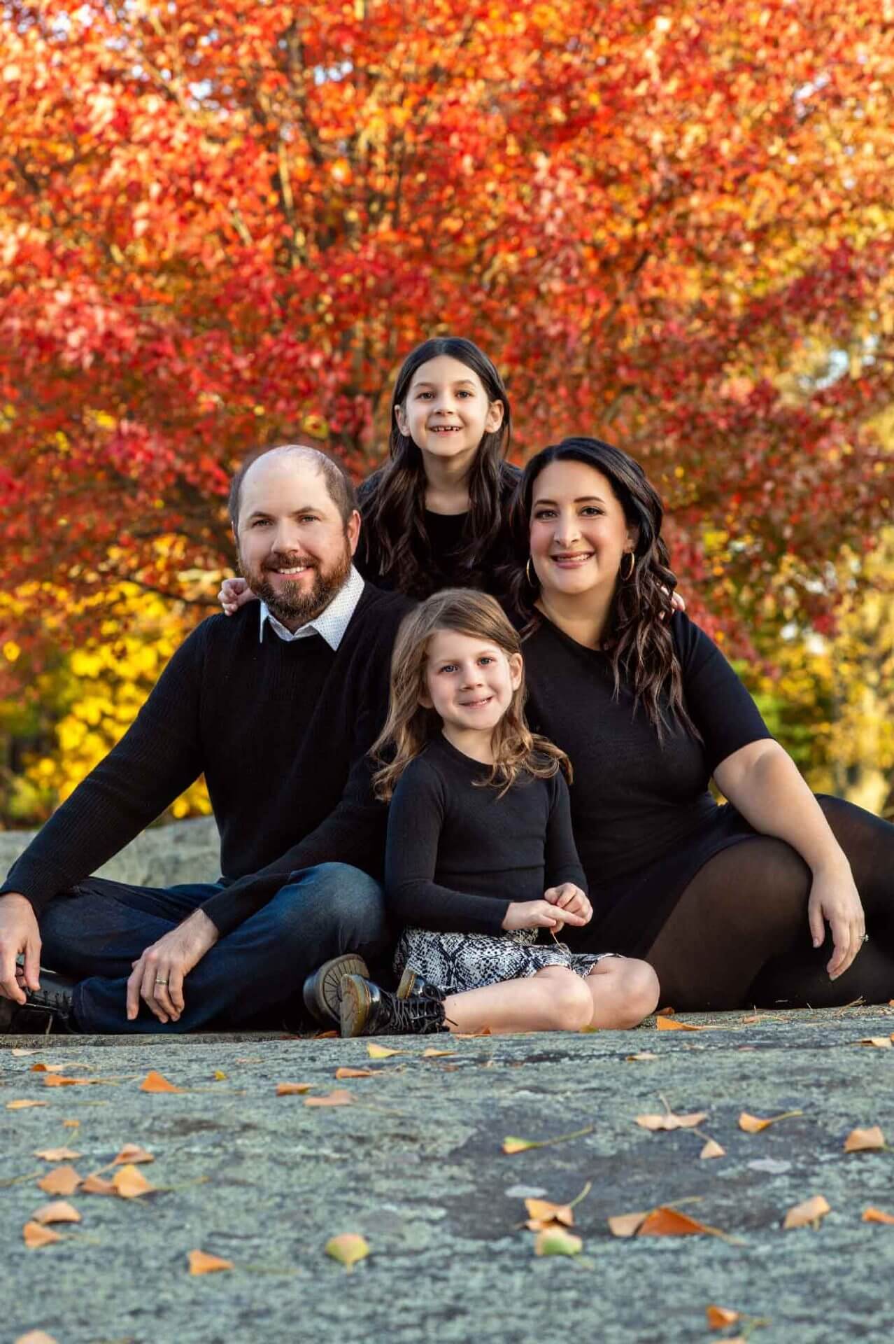 A family of four sits outdoors on a stone surface in front of a tree with bright red autumn leaves, smiling at the camera.
