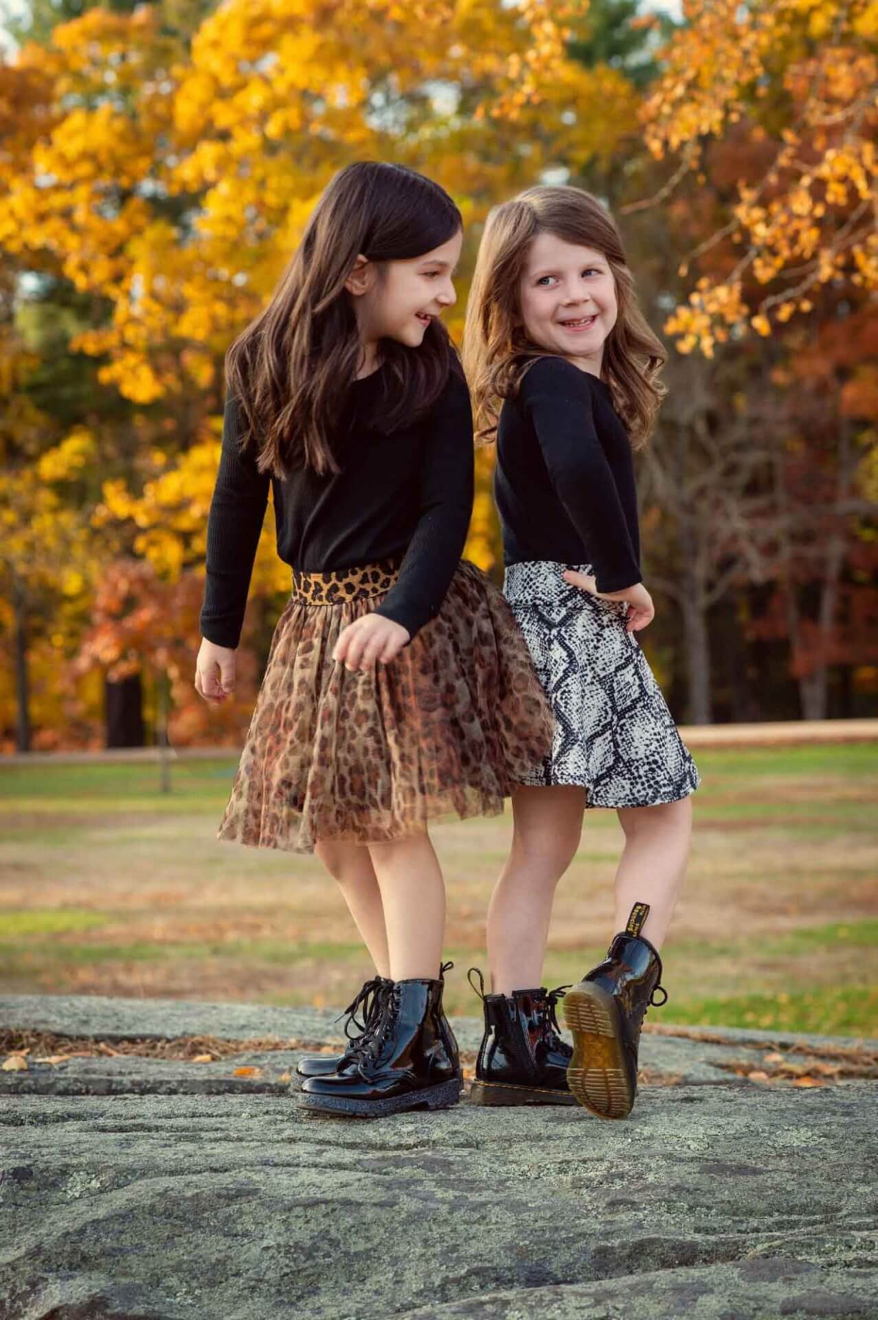 Two young girls stand back to back outdoors in autumn, wearing black long-sleeve tops, patterned skirts, and black boots, smiling with colorful fall leaves in the background.