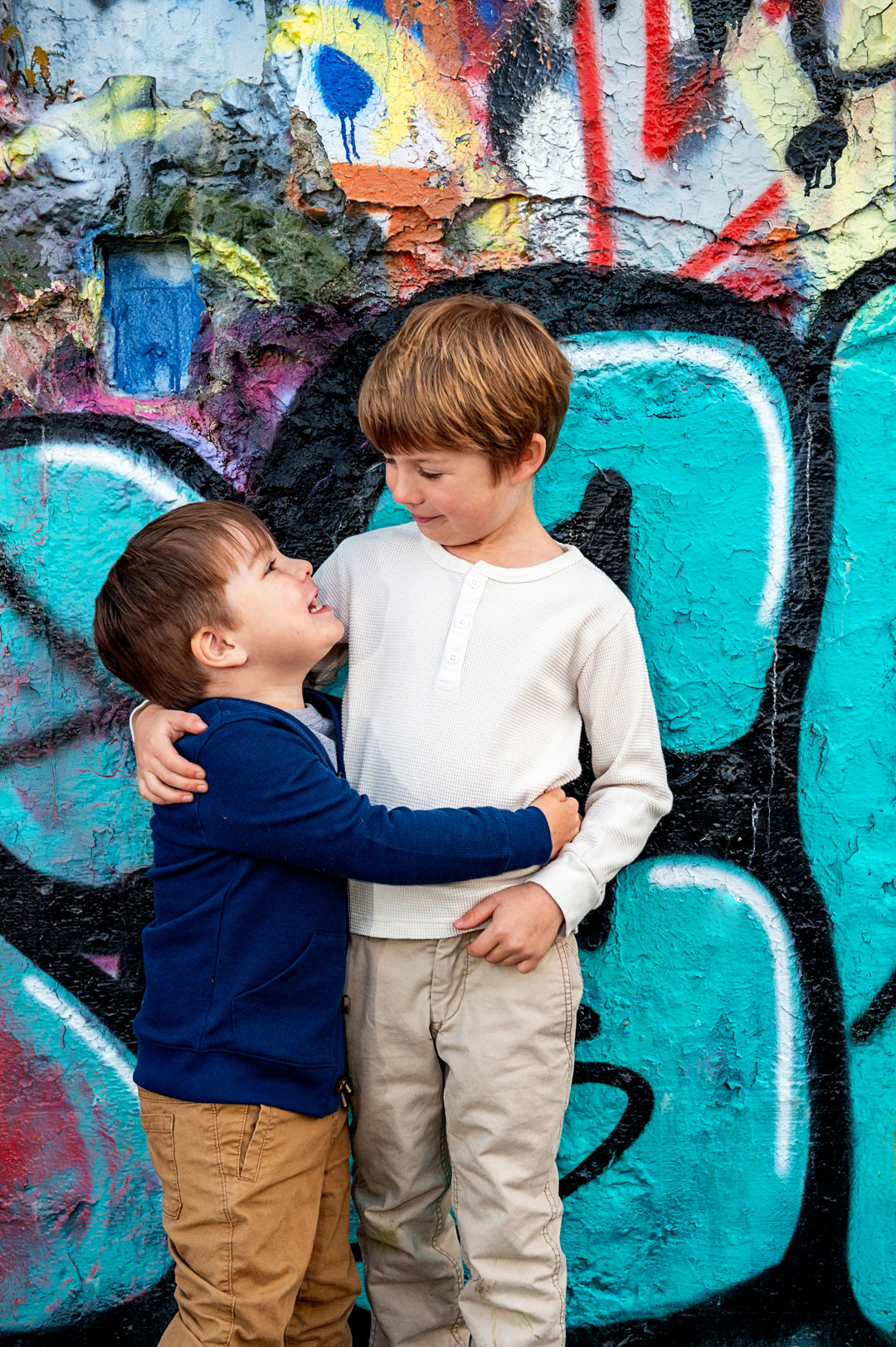 Two young boys standing in front of a colorful graffiti wall, smiling and hugging each other.
