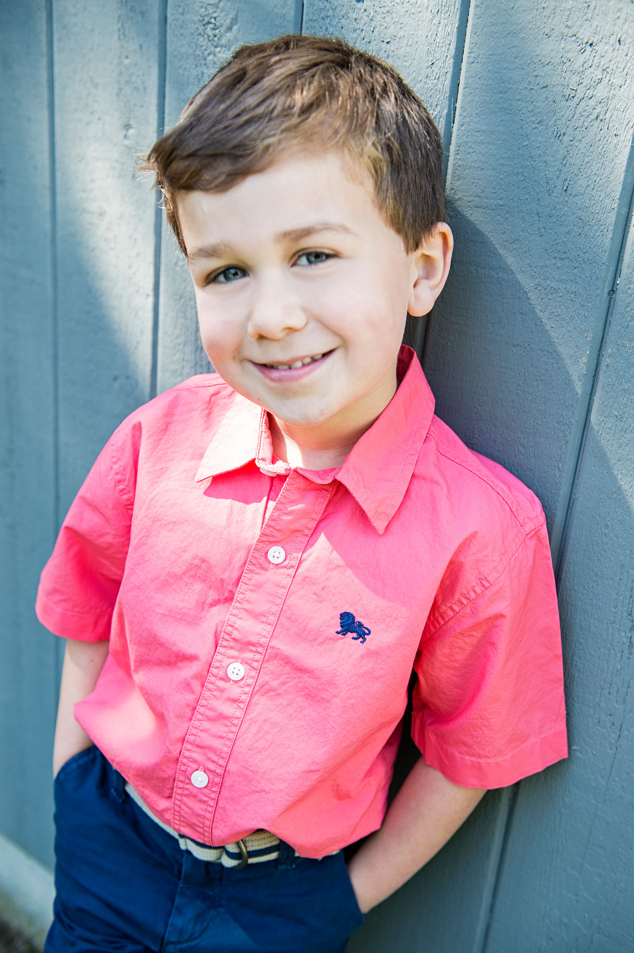 family-portrait-photographer-boy-by-blue-wall Young boy with light brown hair wearing a bright pink button-up shirt and dark blue pants, standing against a gray wooden wall and smiling at the camera.