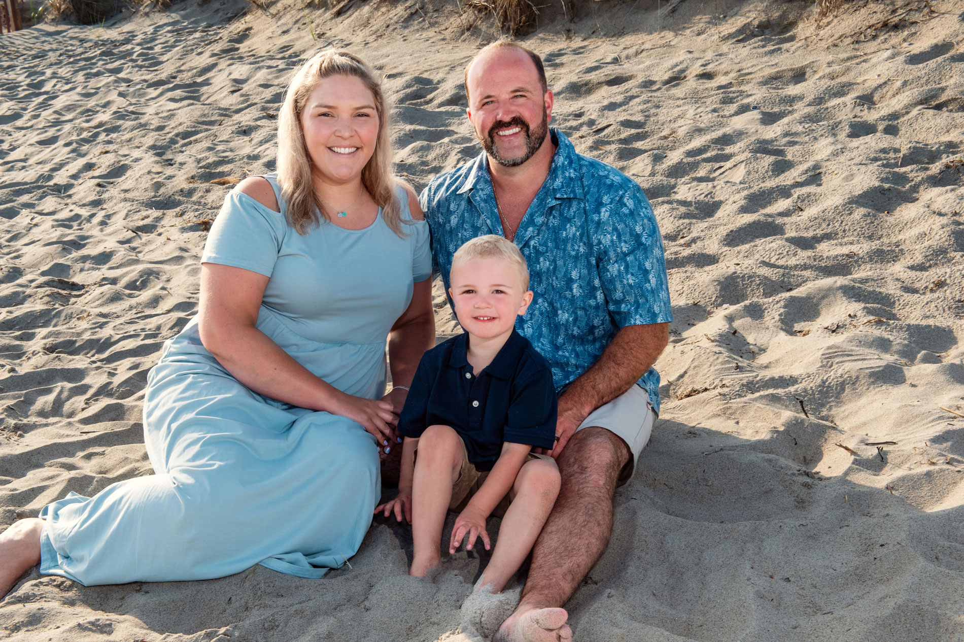family-portrait-photographer-on-rhode-island-beach A woman, a man, and a young boy sit together on sandy beach, smiling at the camera on a sunny day.