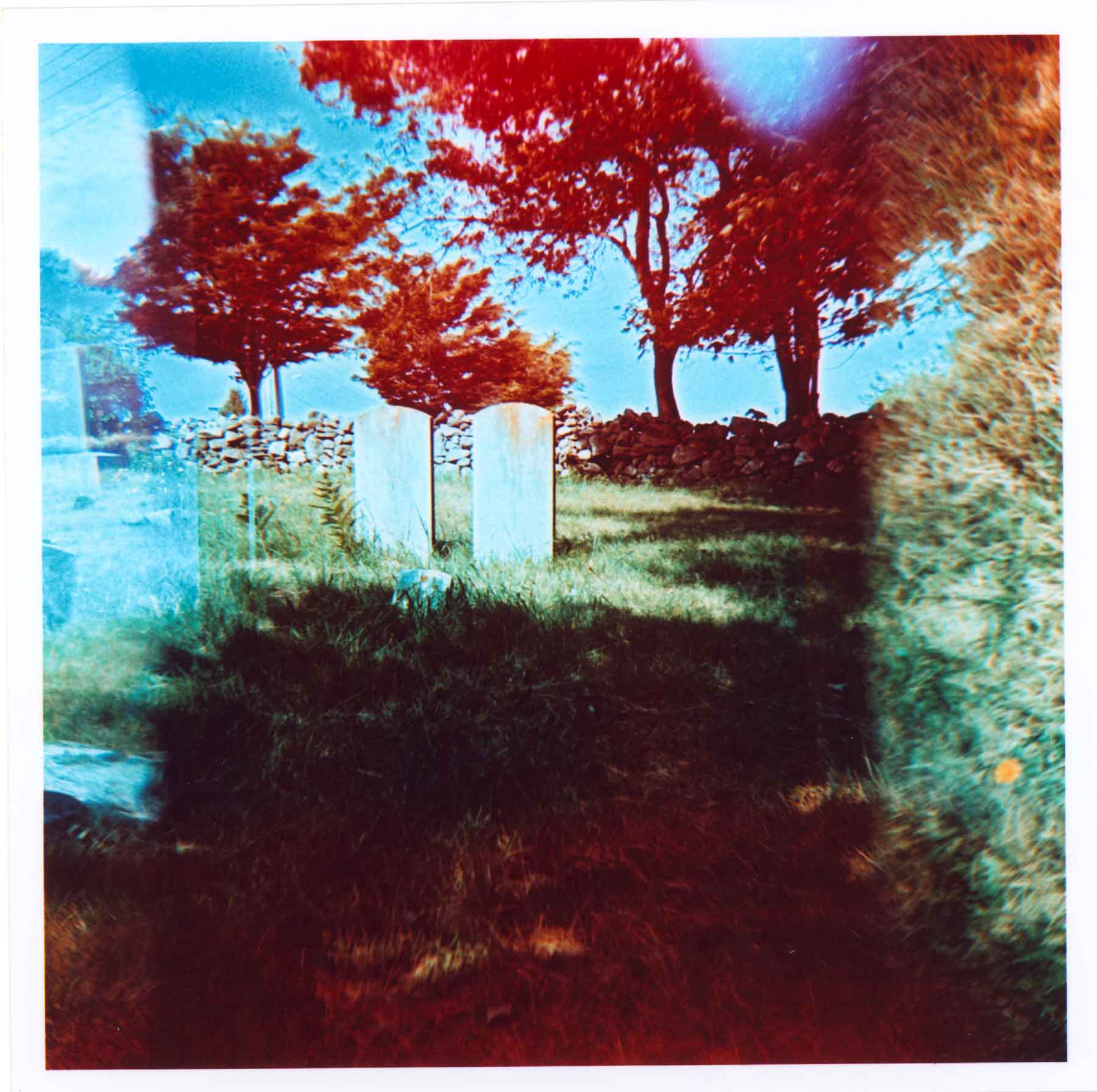 fine-art-photography-peeling-lo-fi-Headstones Two old gravestones stand in a grassy cemetery with trees and a stone wall in the background; the image has a red and blue light leak effect.
