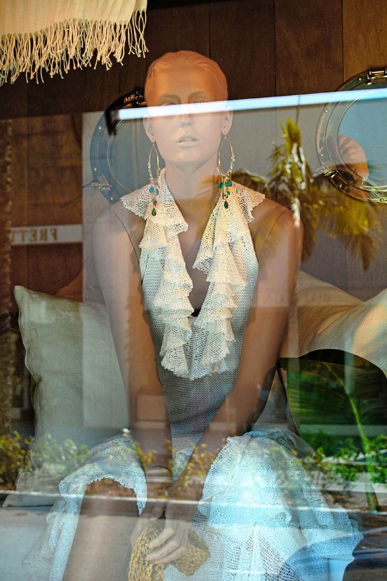 fine-art-photography-reflections-Blind-Shopper A mannequin dressed in a ruffled, sleeveless outfit sits behind a glass window, with reflections of palm trees and outdoor scenery visible on the glass.