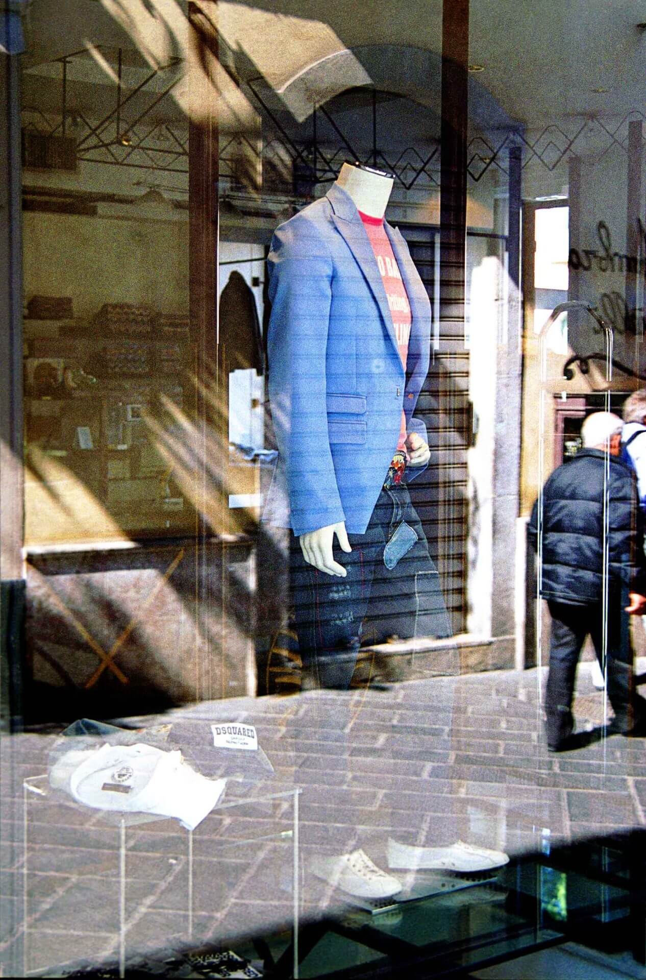 fine-art-photography-reflections-Business-Suit A headless mannequin in a blue blazer and red shirt is displayed in a clothing store window, with reflections of people and buildings on the glass.