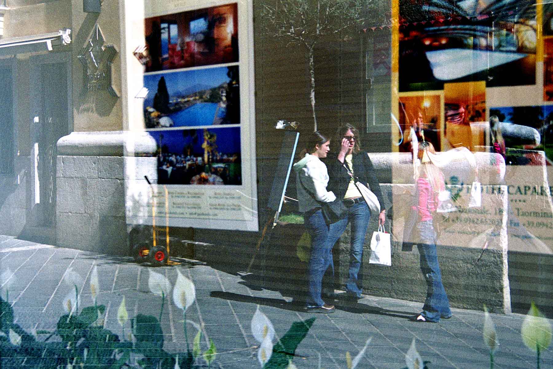 fine-art-photography-reflections-Girls-Shopping Three people stand and talk on a sidewalk, reflected in a window displaying posters and images, with flowers visible in the foreground.