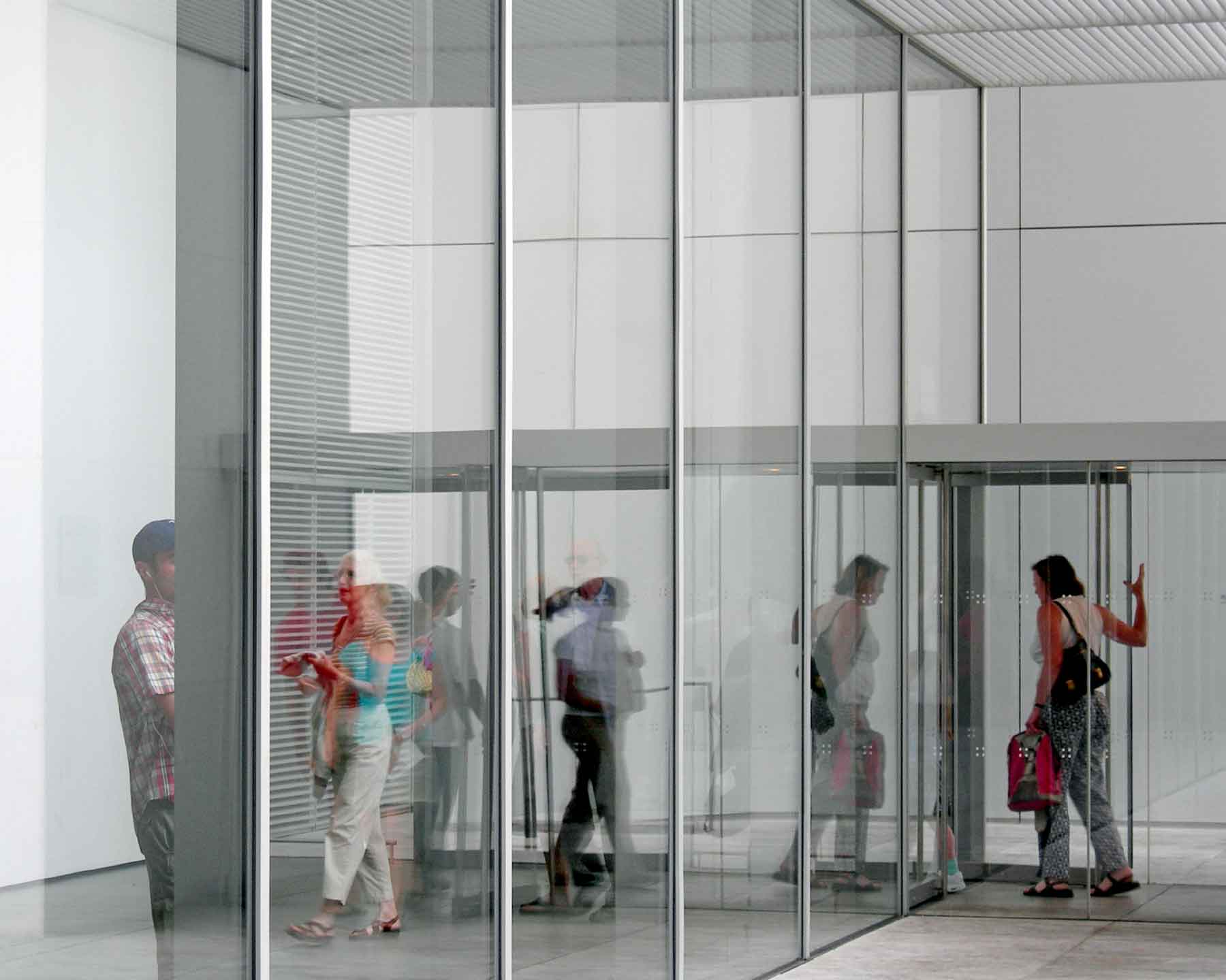 fine-art-photography-reflections-Monotony Several people are seen through large glass panels as they walk through the entrance of a modern building.