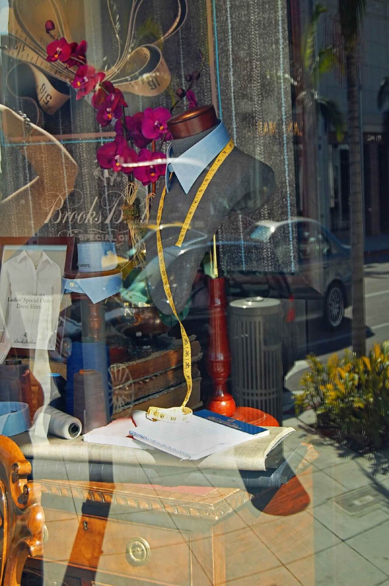 fine-art-photography-reflections-Tailored A tailor’s mannequin with a shirt and measuring tape is displayed in a shop window, surrounded by fabric, tailoring tools, and reflections of the street outside.