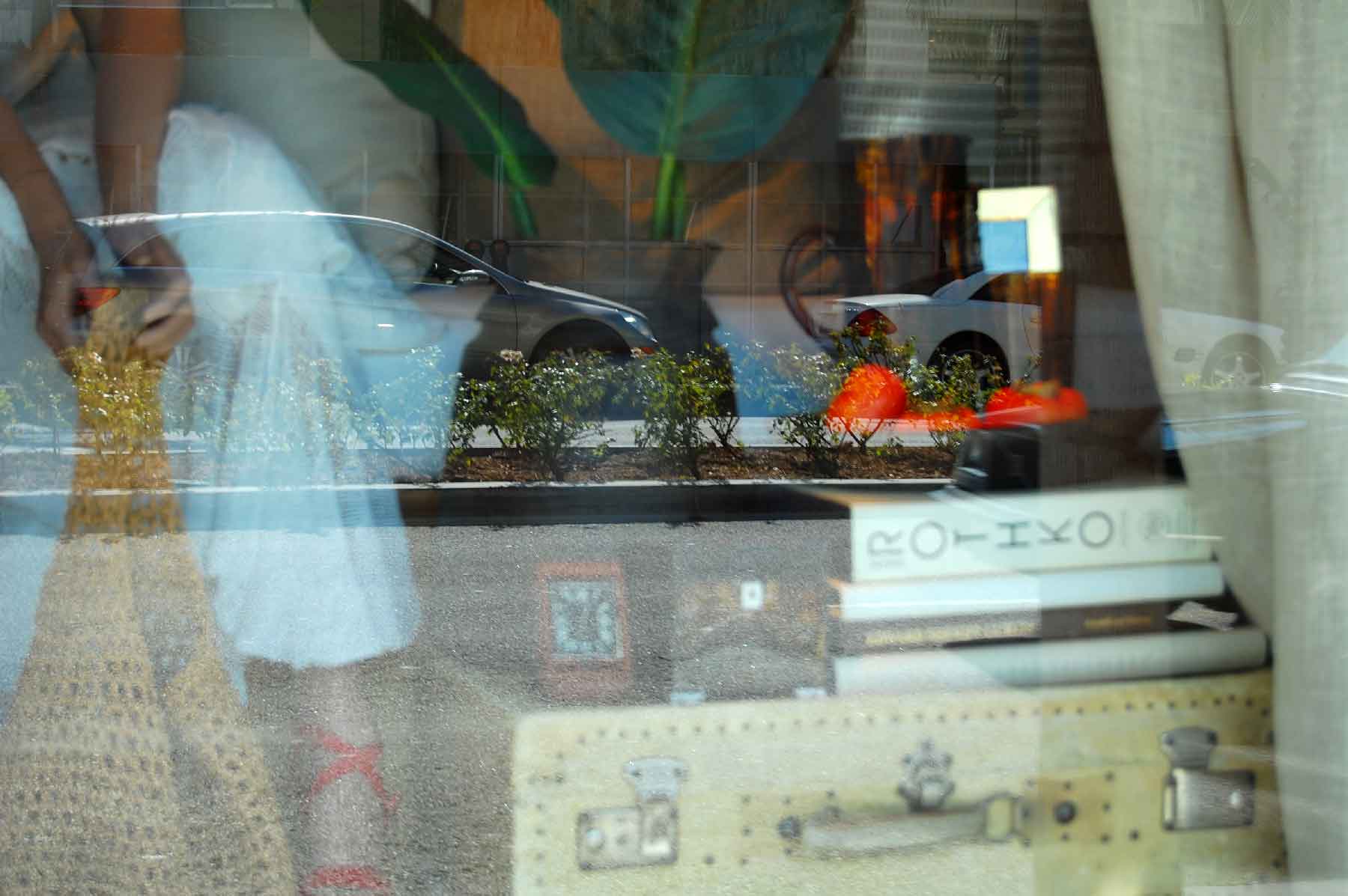 fine-art-photography-reflections-Waiting for Rothko A reflection in a window shows a parked car, greenery, and indoor objects like books, a basket, and a suitcase, with a person partially visible on the left.