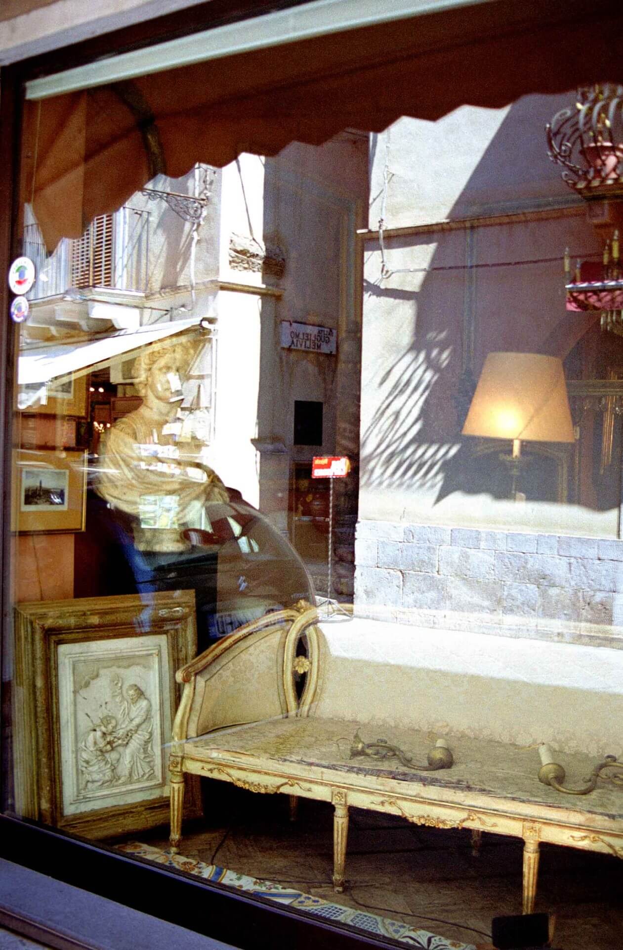 fine-art-photography-reflections-couch A storefront window displays an antique settee, framed artwork, and a sculpture, with street reflections visible in the glass.