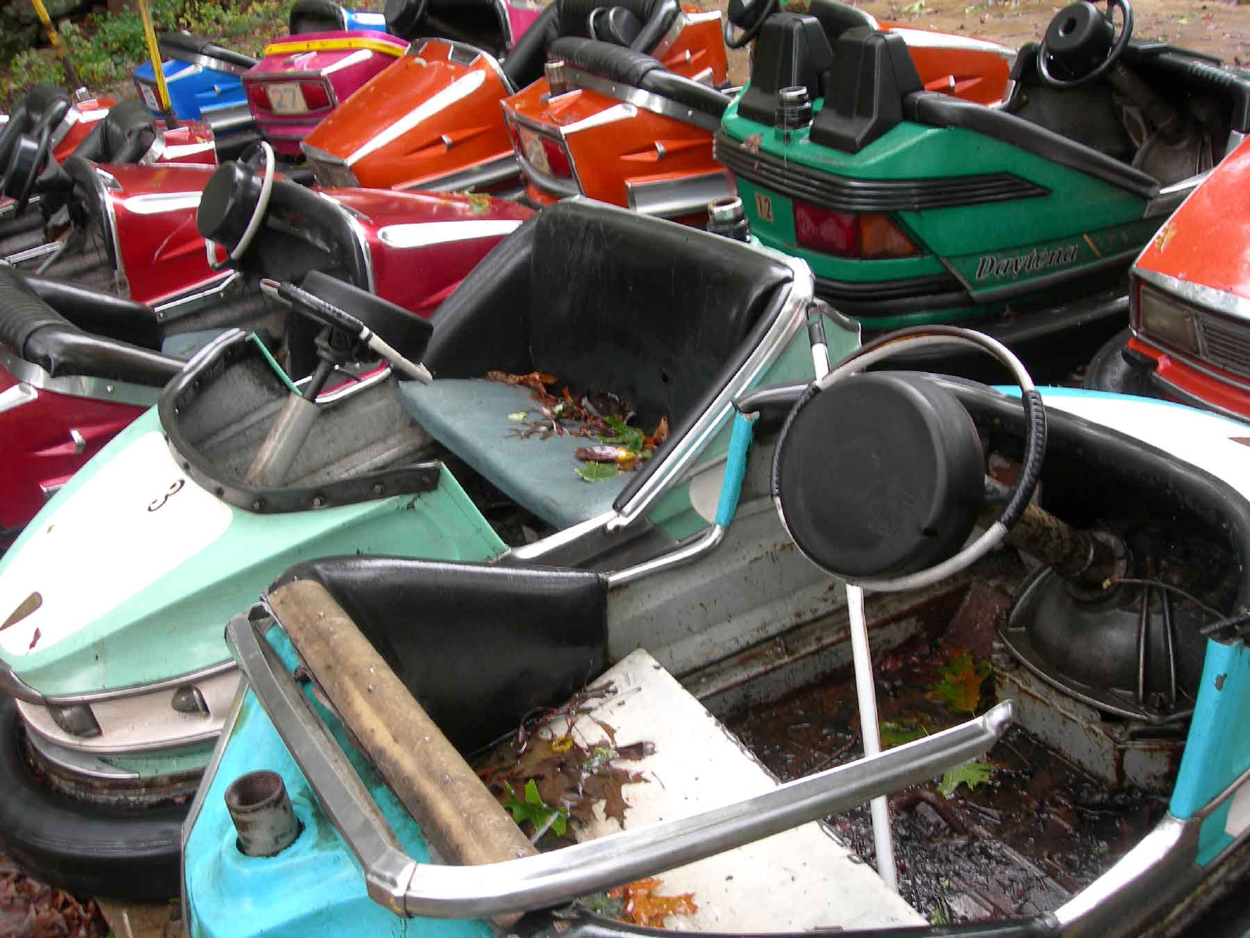 Old bumper cars with faded paint and leaves inside are crowded together outdoors, showing signs of rust and disuse.